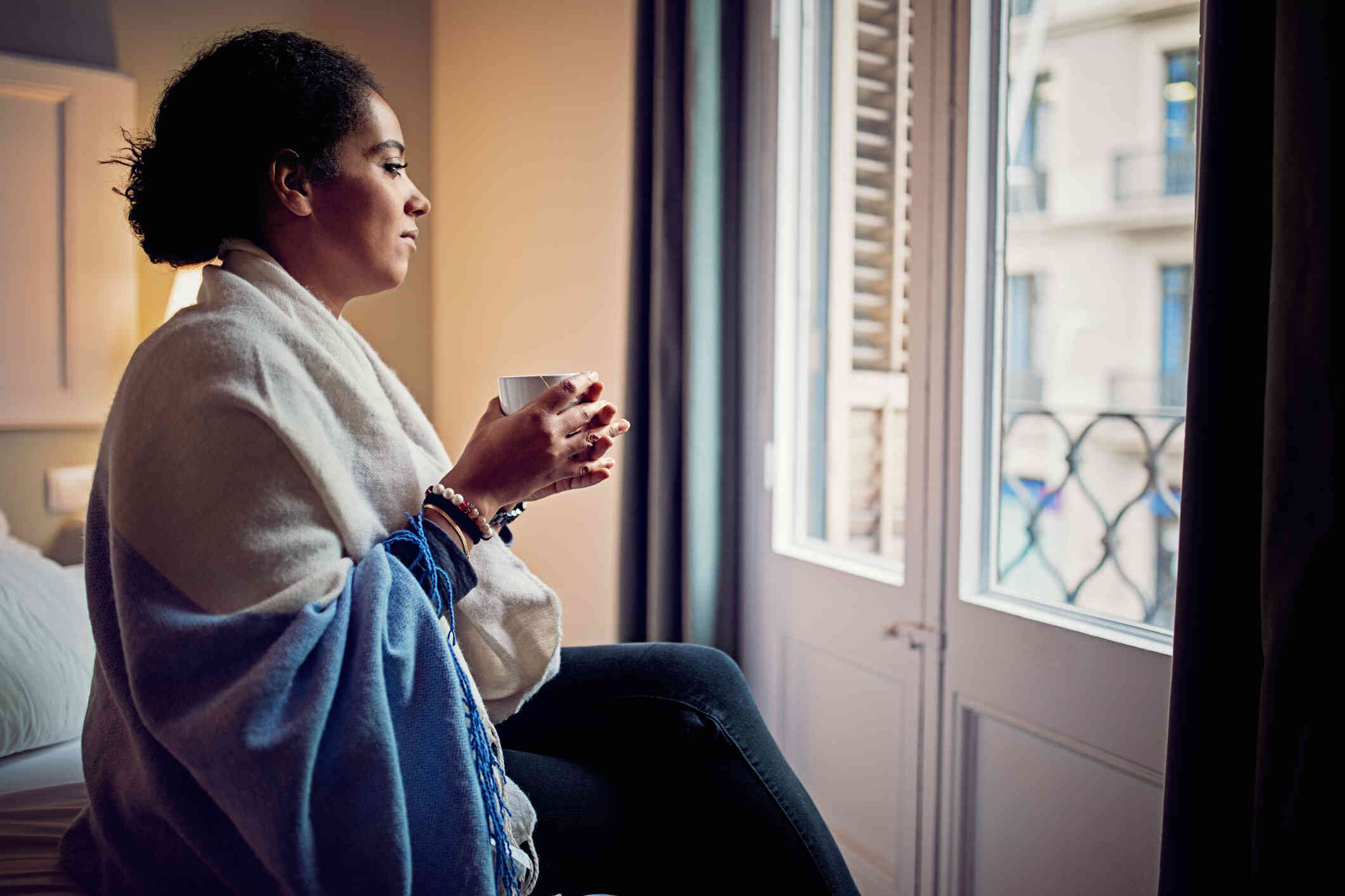 A woman in a large scarf holds a mug in her hands while gazing out of a window with a sad expression.